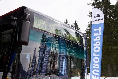 Auf einem Bus steht Skadi Loppet als Ziel. In der Winschutzscheibe spiegelt sich die verschneite Landschaft sowie Euro Loppet Fahnen. | © Bodenmais Tourismus & Marketing GmbH